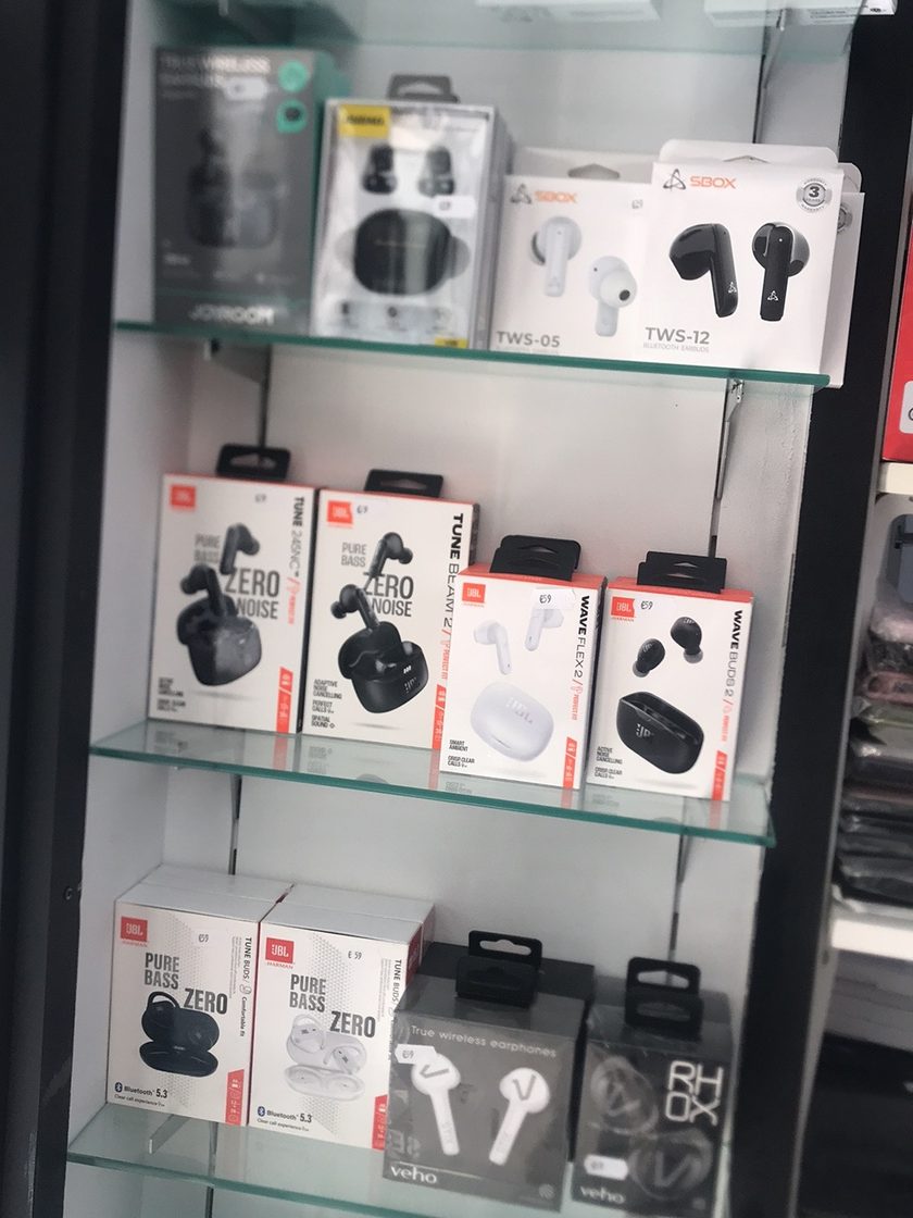 A glass cabinet of boxed wireless earbuds, JBL, SBOX, Joyroom and Veho, with handwritten euro price tags on the shelf edges